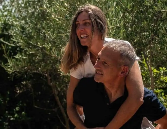 The hosts of 4U Bed&Spa smiling, she riding on his back, in front of an olive tree