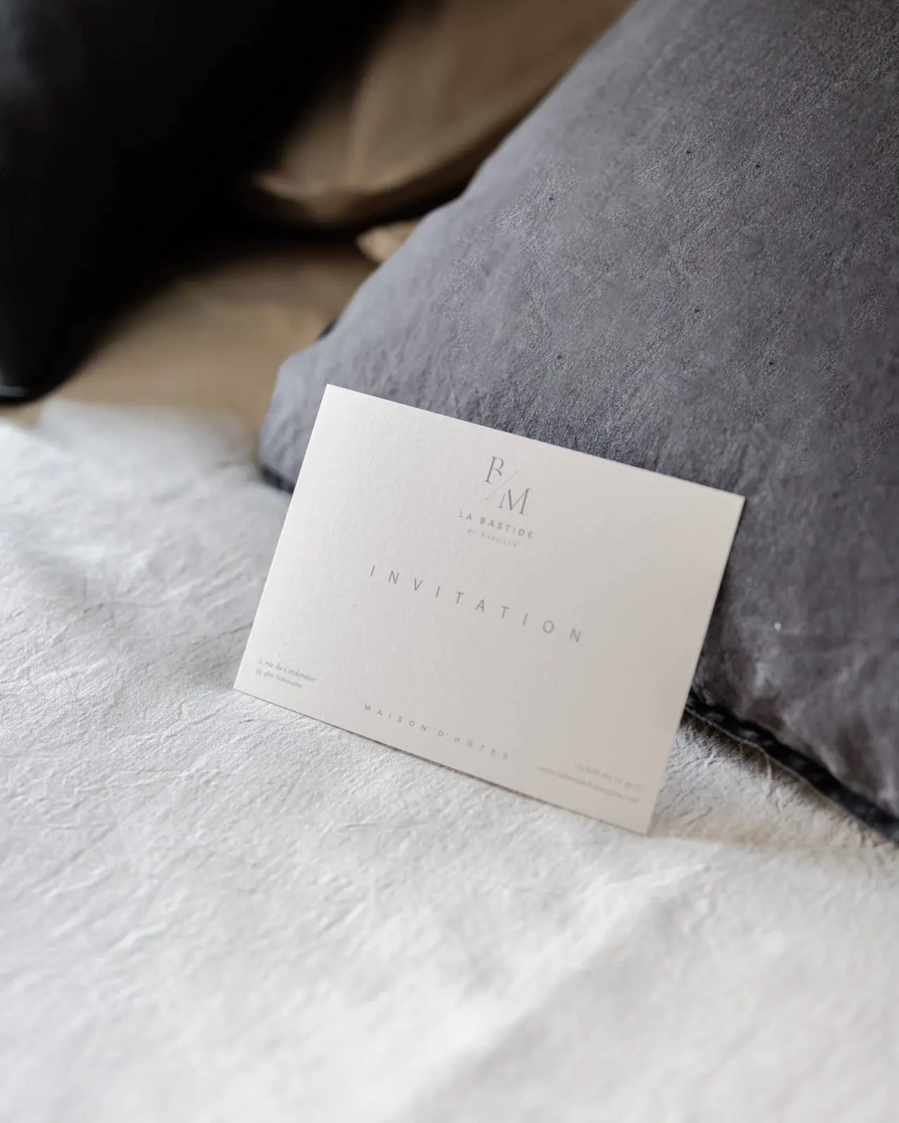 La Bastide de Margaux invitation card propped against a grey linen cushion on a white bed