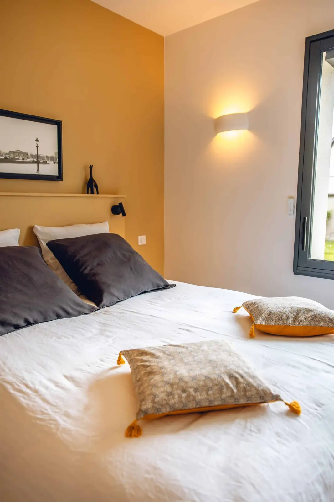 Ochre-walled bedroom at Bread et Couette with grey cushions, tasselled pillows and a black-and-white photo above the bed