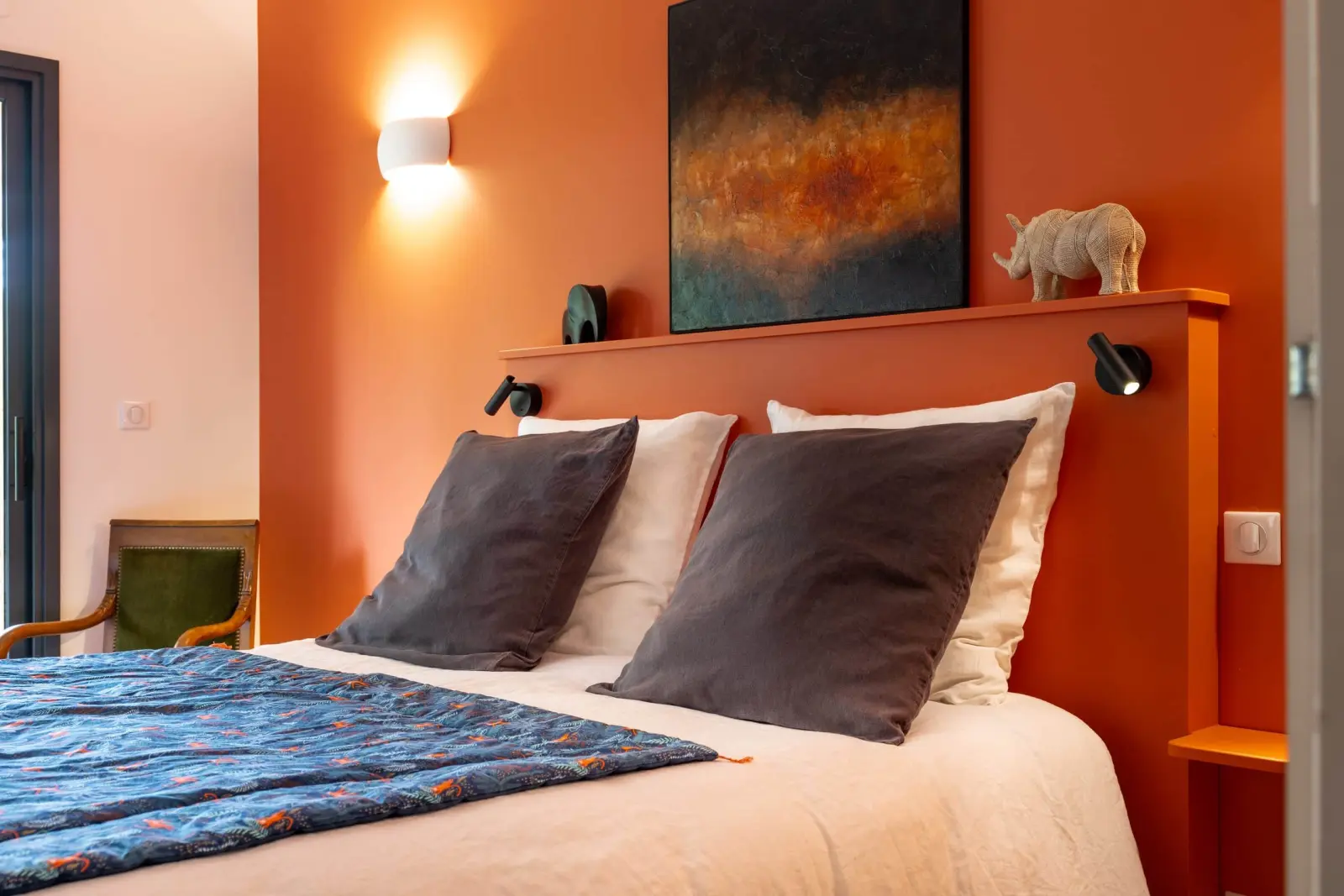 Orange-walled bedroom at Bread et Couette with grey cushions, an abstract painting and a rhino figure above the bed