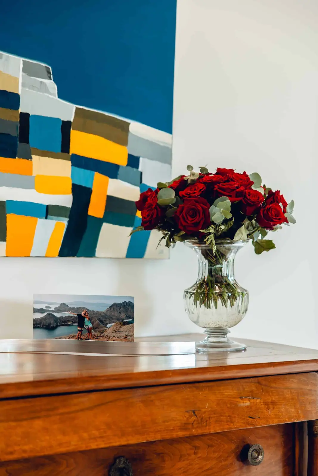 Wooden chest of drawers with red roses in a glass vase and a colourful painting, at Bread et Couette