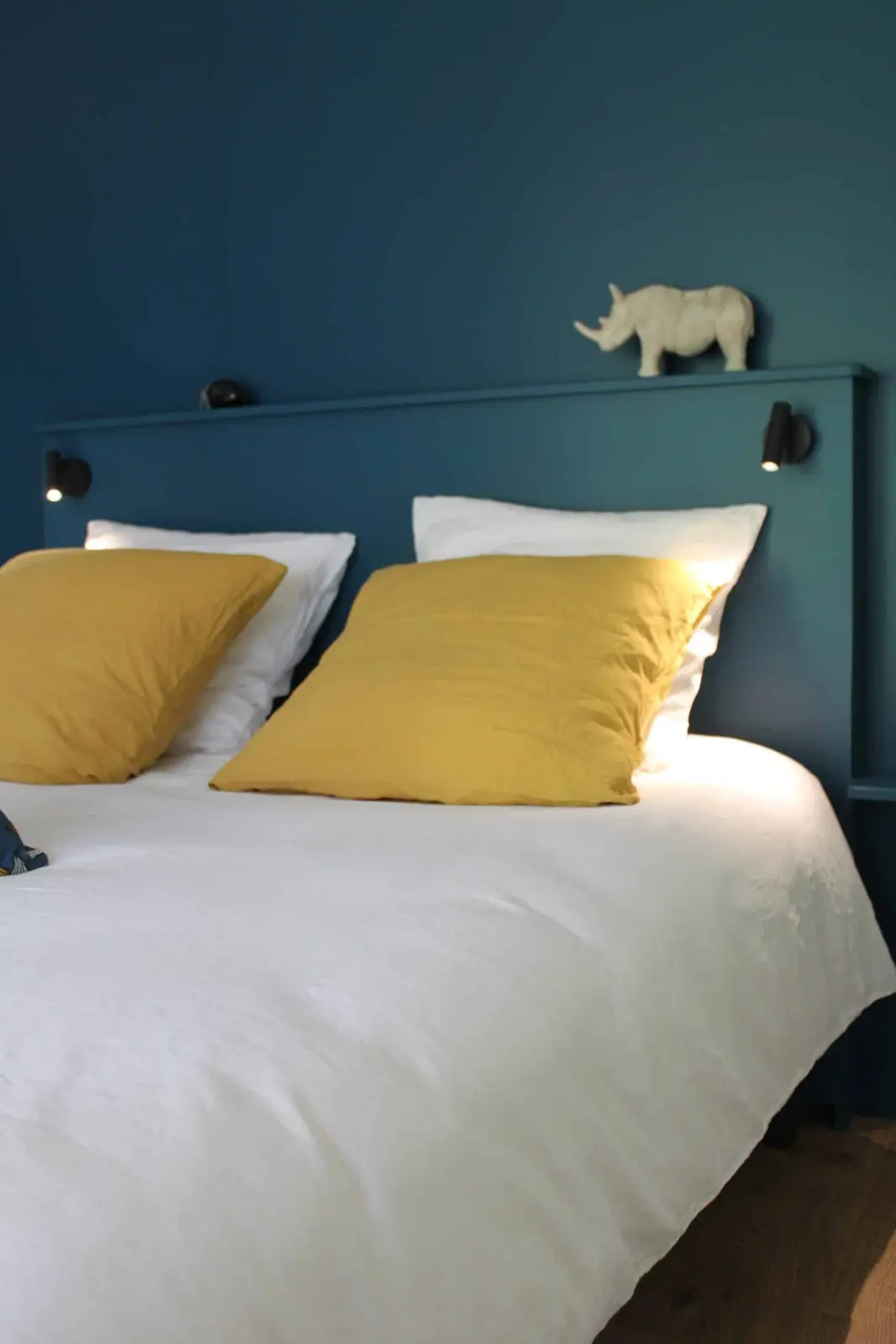 Bed with mustard-yellow cushions and white linen against a teal headboard topped by a rhino figure, at Bread et Couette