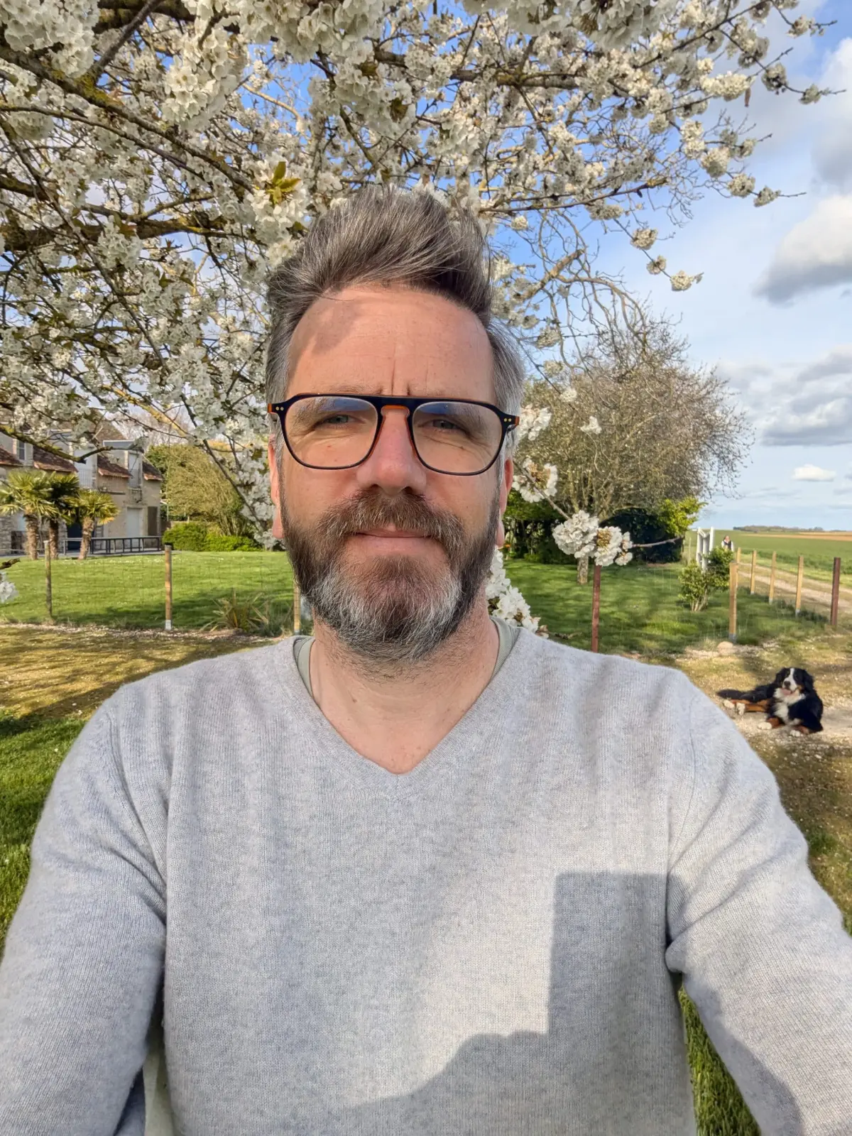 The host of Domaine de Champouteau, glasses and grey jumper, under a blossoming cherry tree, a dog lying behind