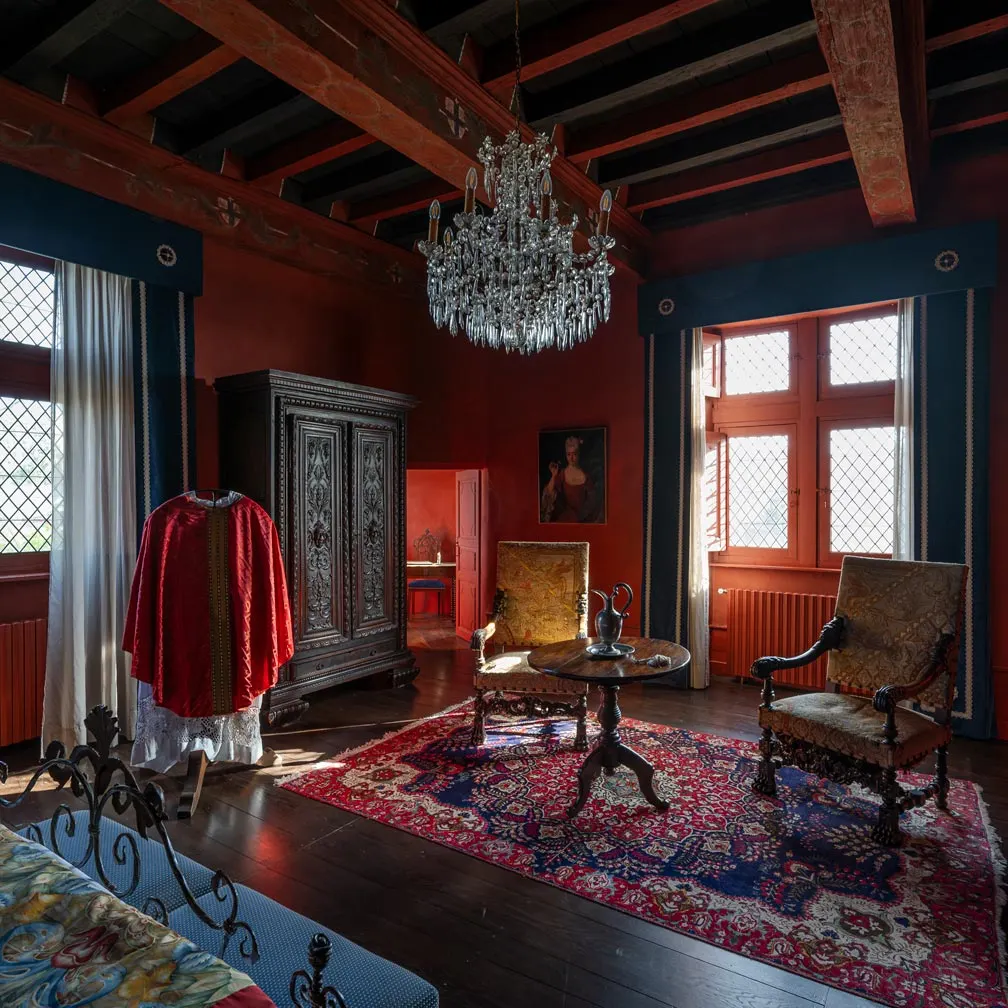 Red-walled room at Château de Canac with beamed ceiling, crystal chandelier, carved wardrobe and antique rug
