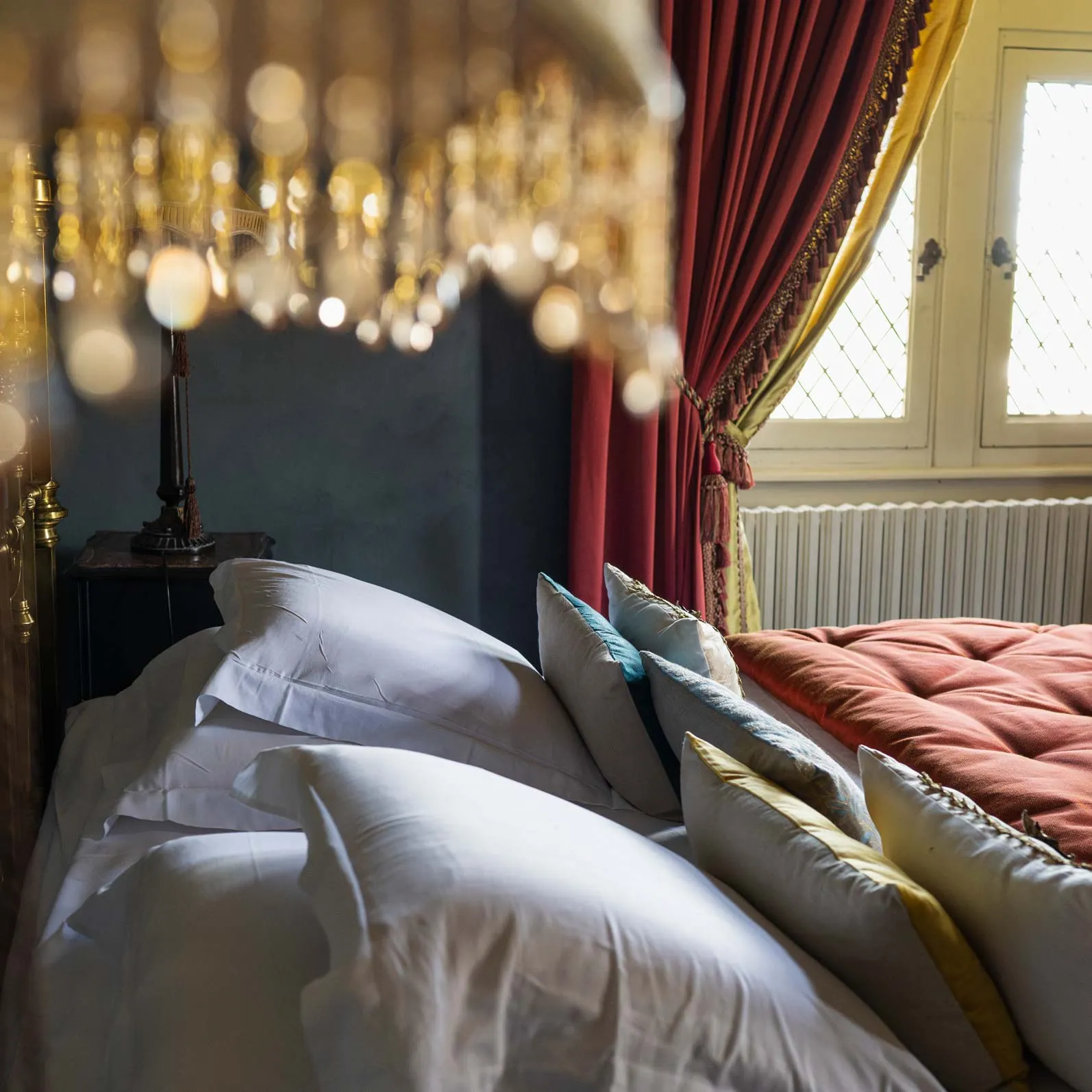 Close-up of a bed with white pillows beneath chandelier droplets and red curtains, at Château de Canac