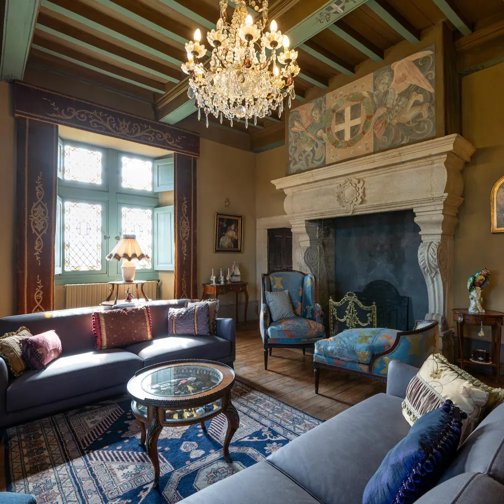 Lounge at Château de Canac with a monumental stone fireplace, crystal chandelier, grey sofas and leaded windows