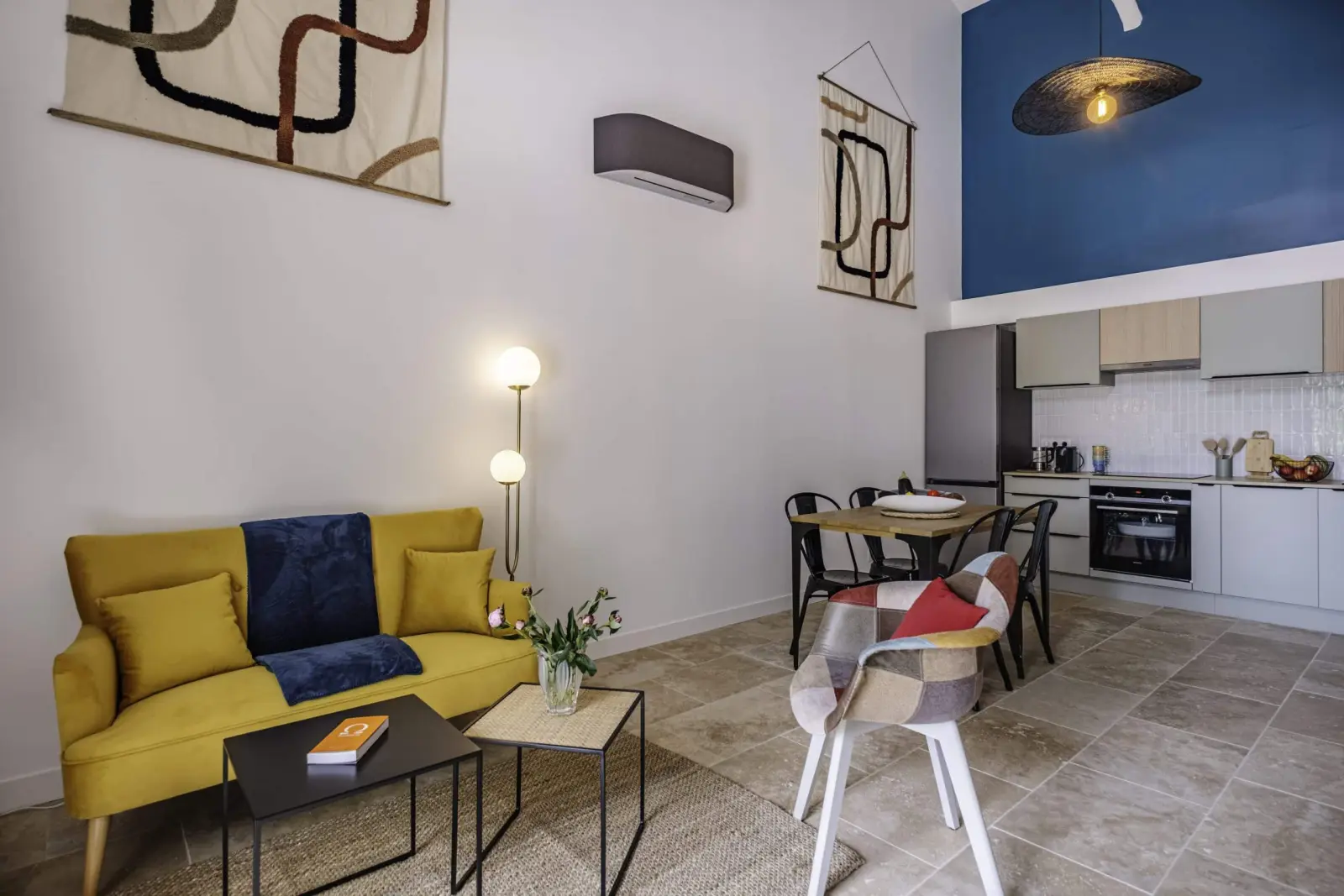 Living room in the Clos Marcamps duplex with yellow sofa, blue wall and open kitchen on a stone floor