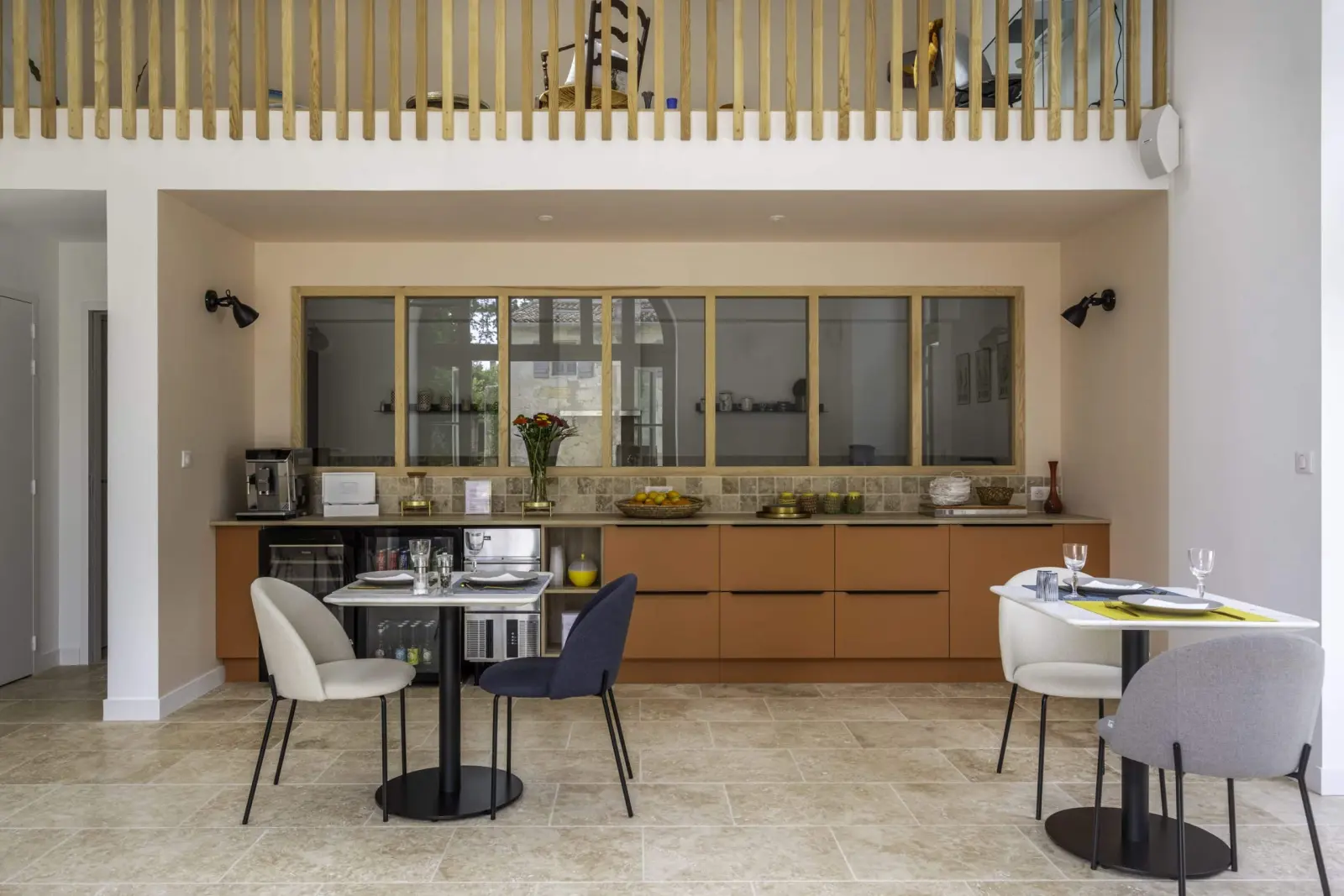 Shared room at Clos Marcamps with orange kitchen units below an interior window, mezzanine and bistro tables