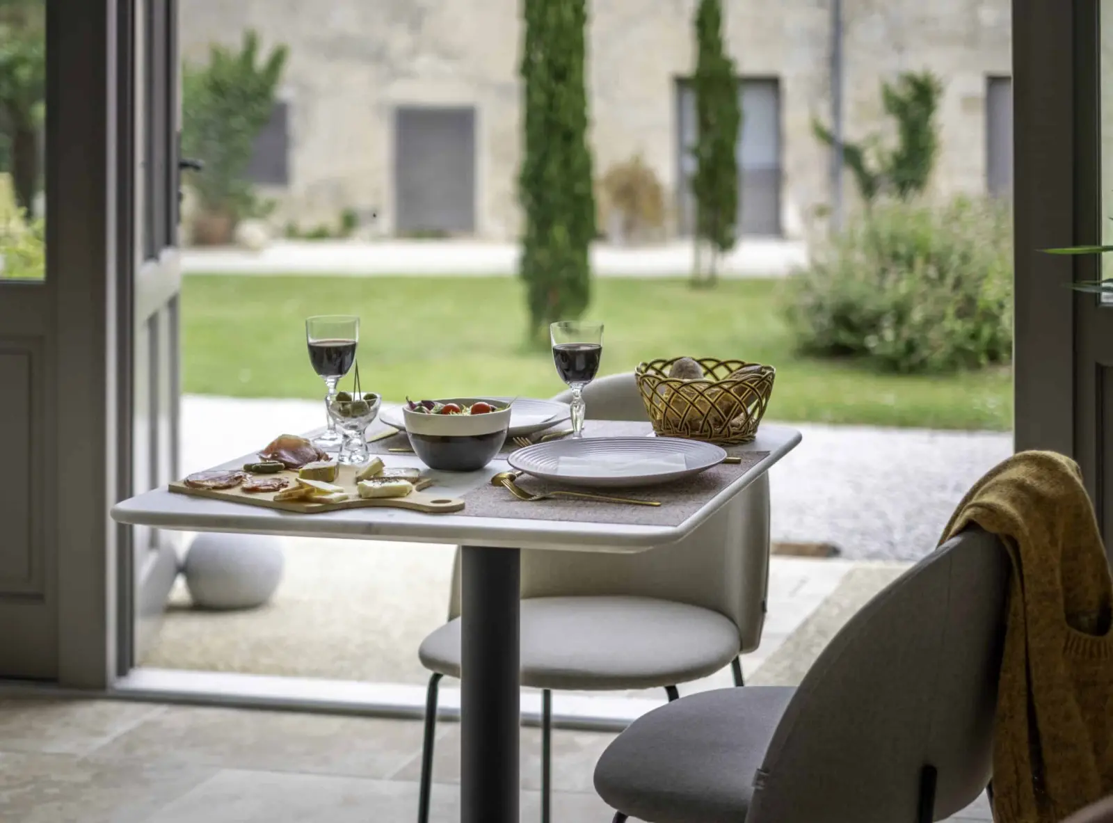Table for two with red wine and a sharing board by a French door open onto the garden, at Clos Marcamps