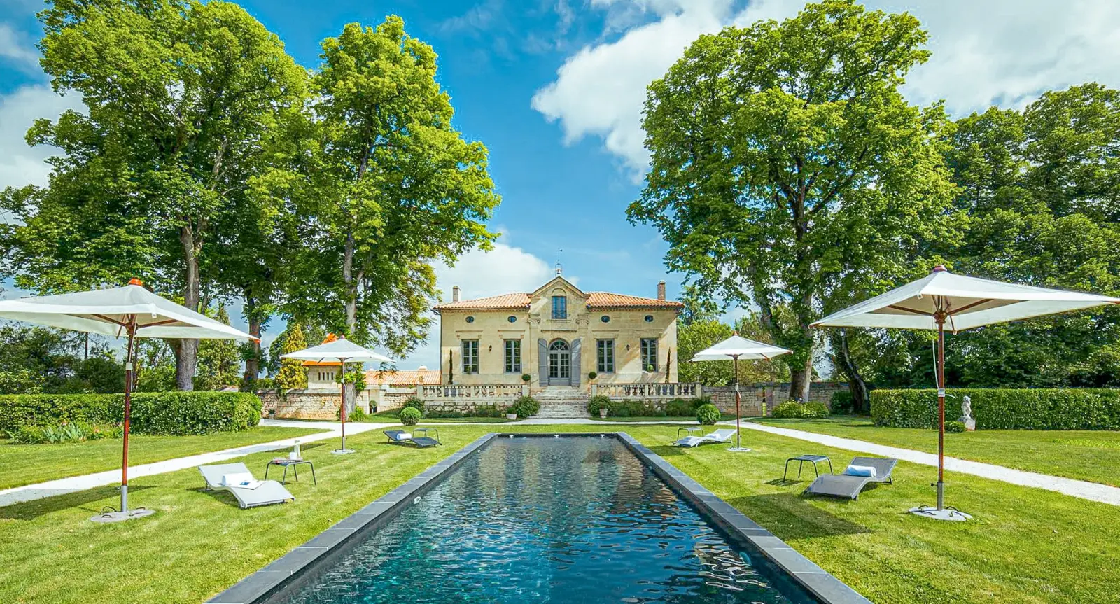 Longue piscine bordée de pelouse et de parasols blancs face à une demeure en pierre sous les arbres