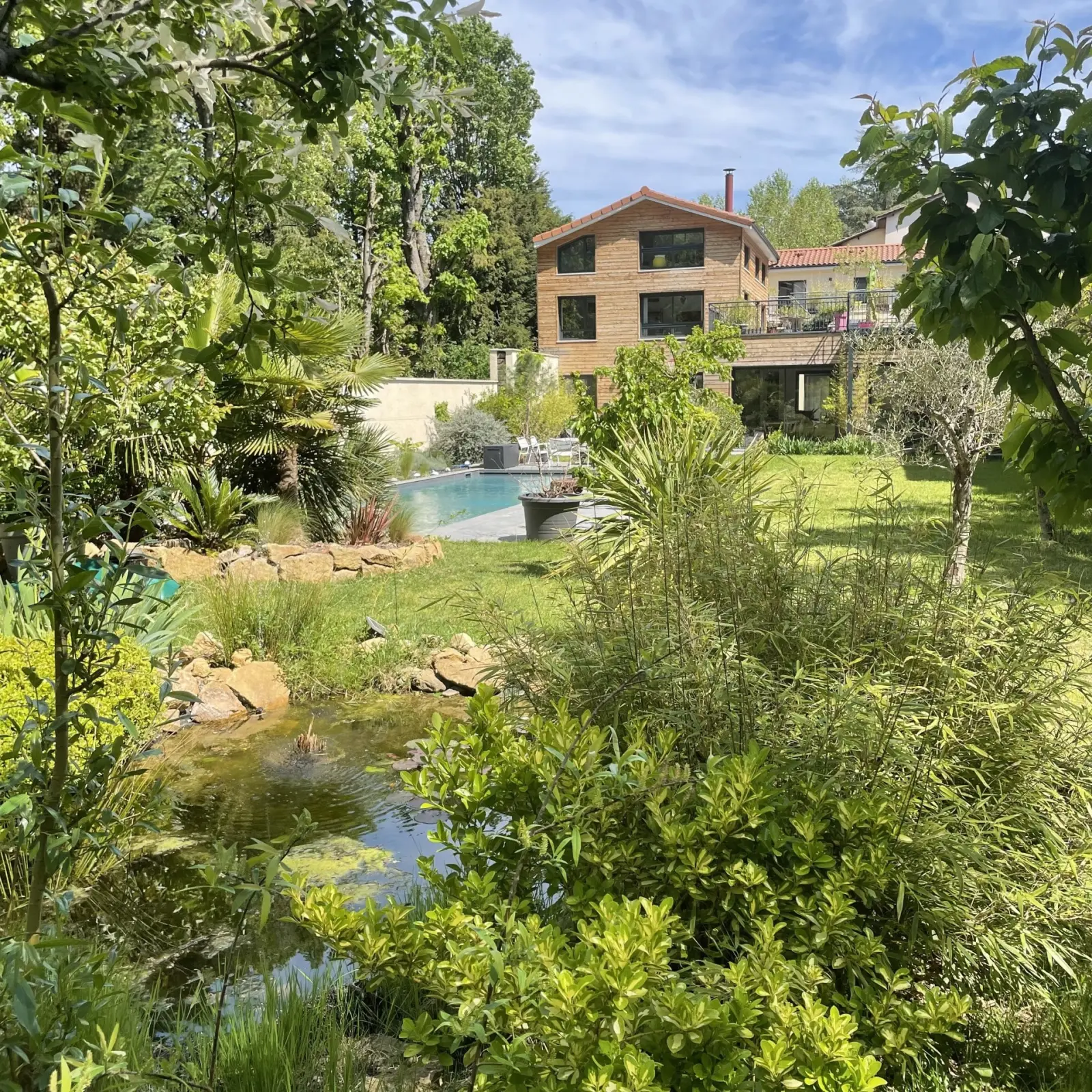 Effet Lodge garden with a pond and greenery, the pool and timber and brick house in the background