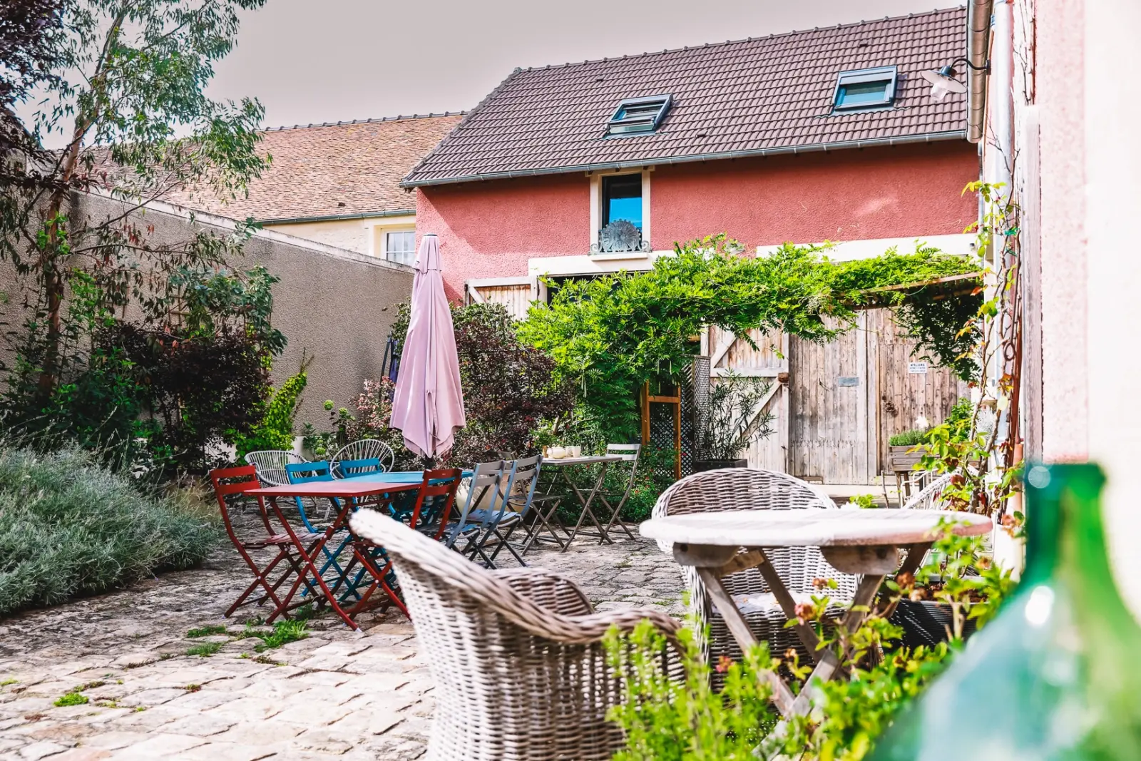 Cour pavée de L'Escale Bellifontaine avec tables colorées, fauteuils en rotin et façade rose