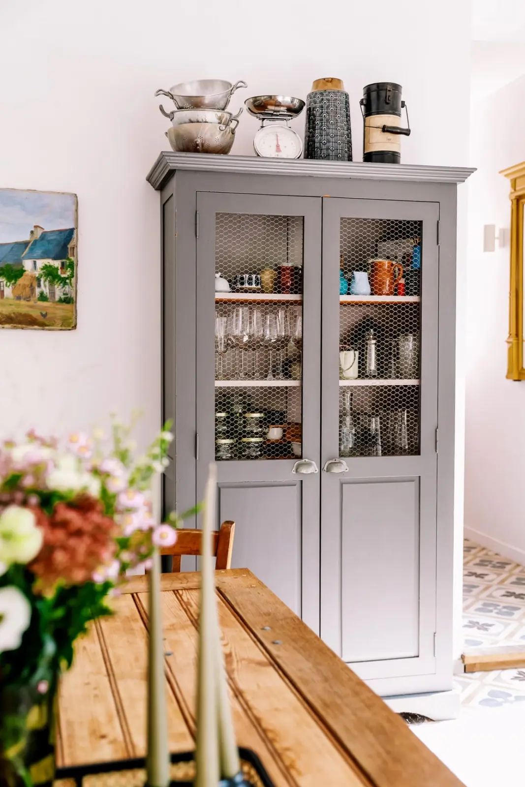 Grey dresser with wire-mesh doors and a wooden table with flowers in the L'escale Bellifontaine kitchen
