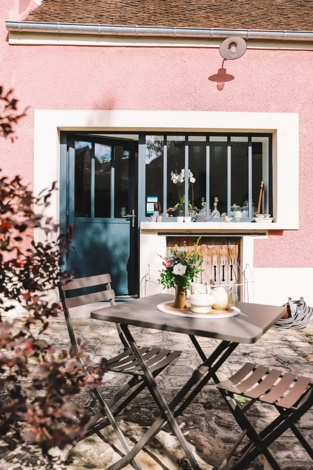 Garden table and folding chairs in front of the pink façade and steel-framed glazing of L'escale Bellifontaine