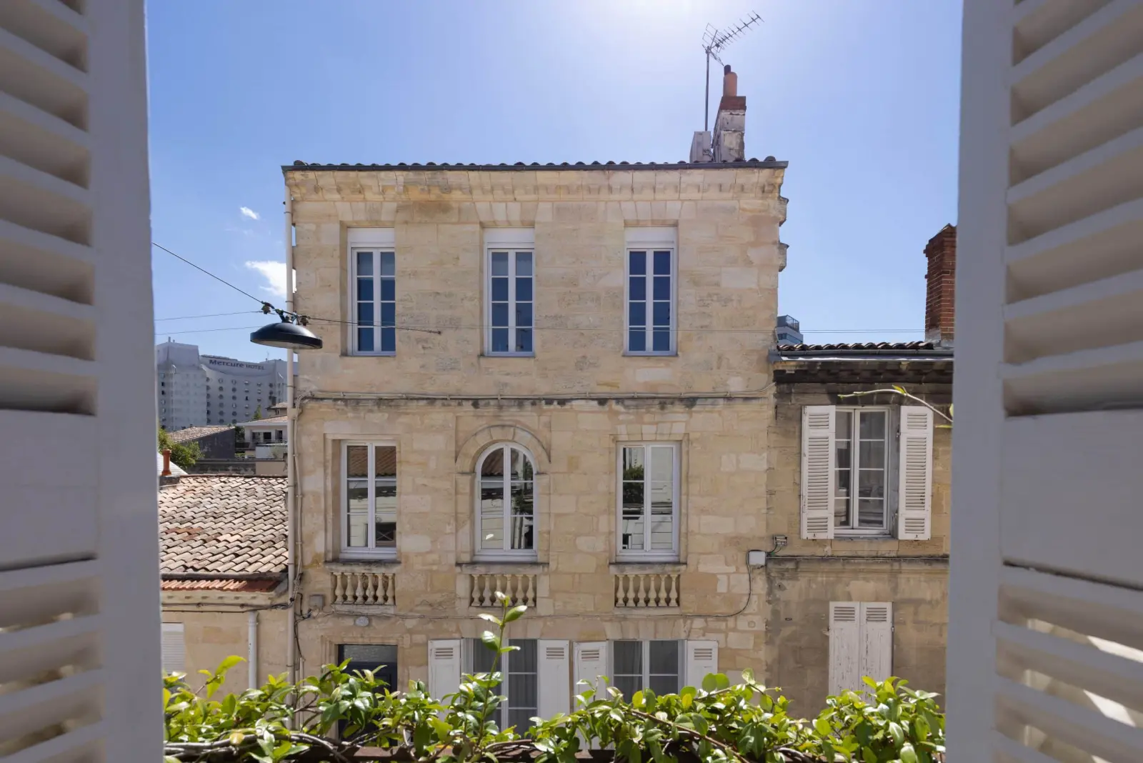 Immeuble en pierre blonde aux balcons en fer forgé, vu entre les volets ouverts d'une fenêtre