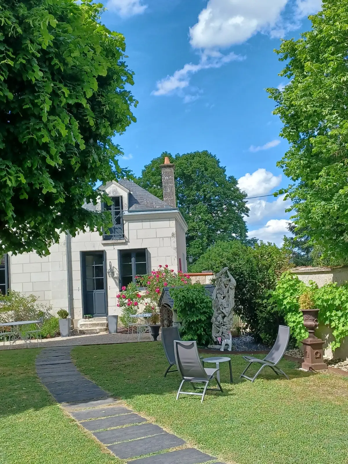 Paved path across the lawn towards a stone house, with loungers and a rose bush in bloom, La Folie Saint Julien