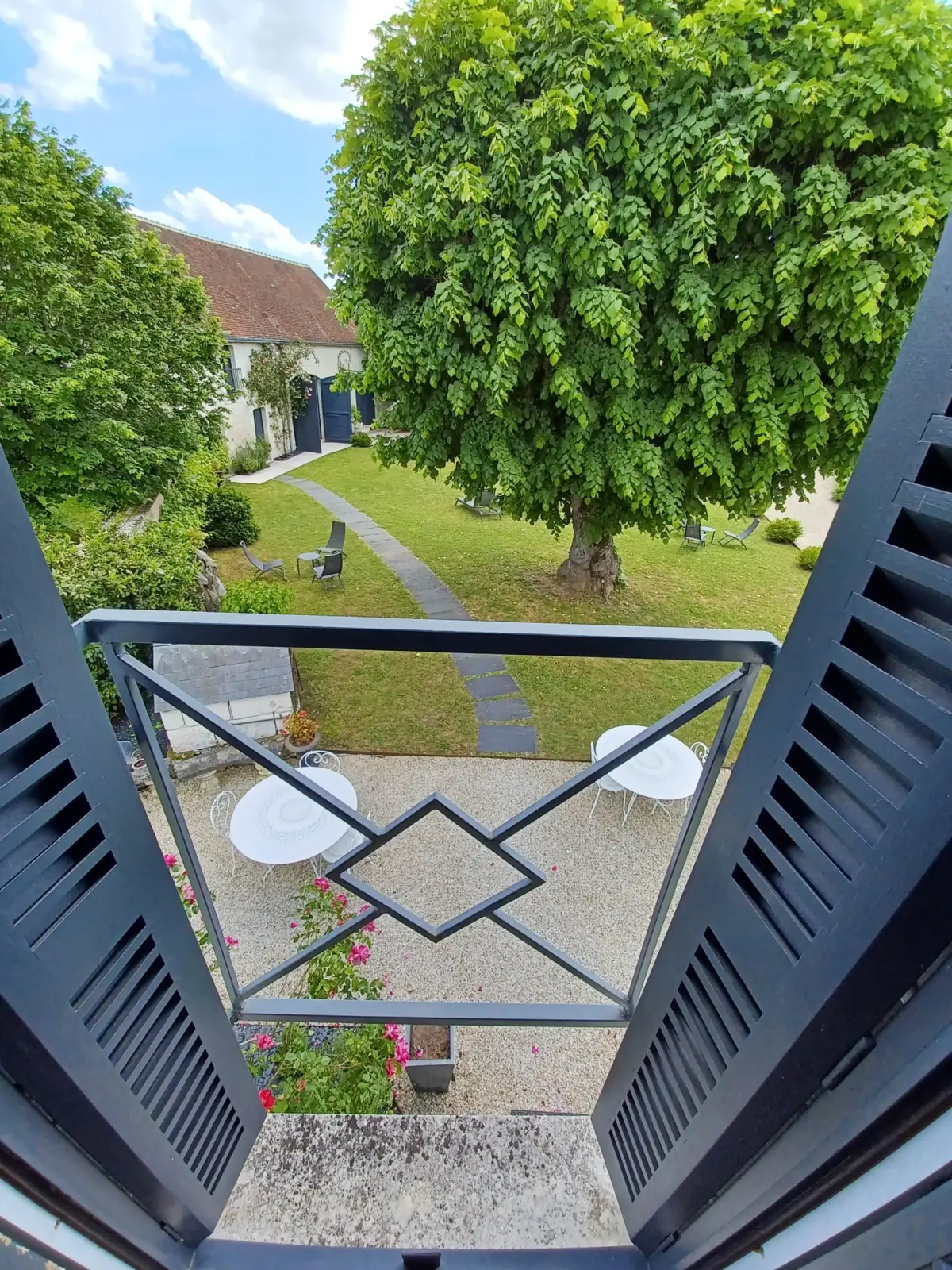 View from a balcony with grey shutters onto the large tree, the lawn and white tables, La Folie Saint Julien