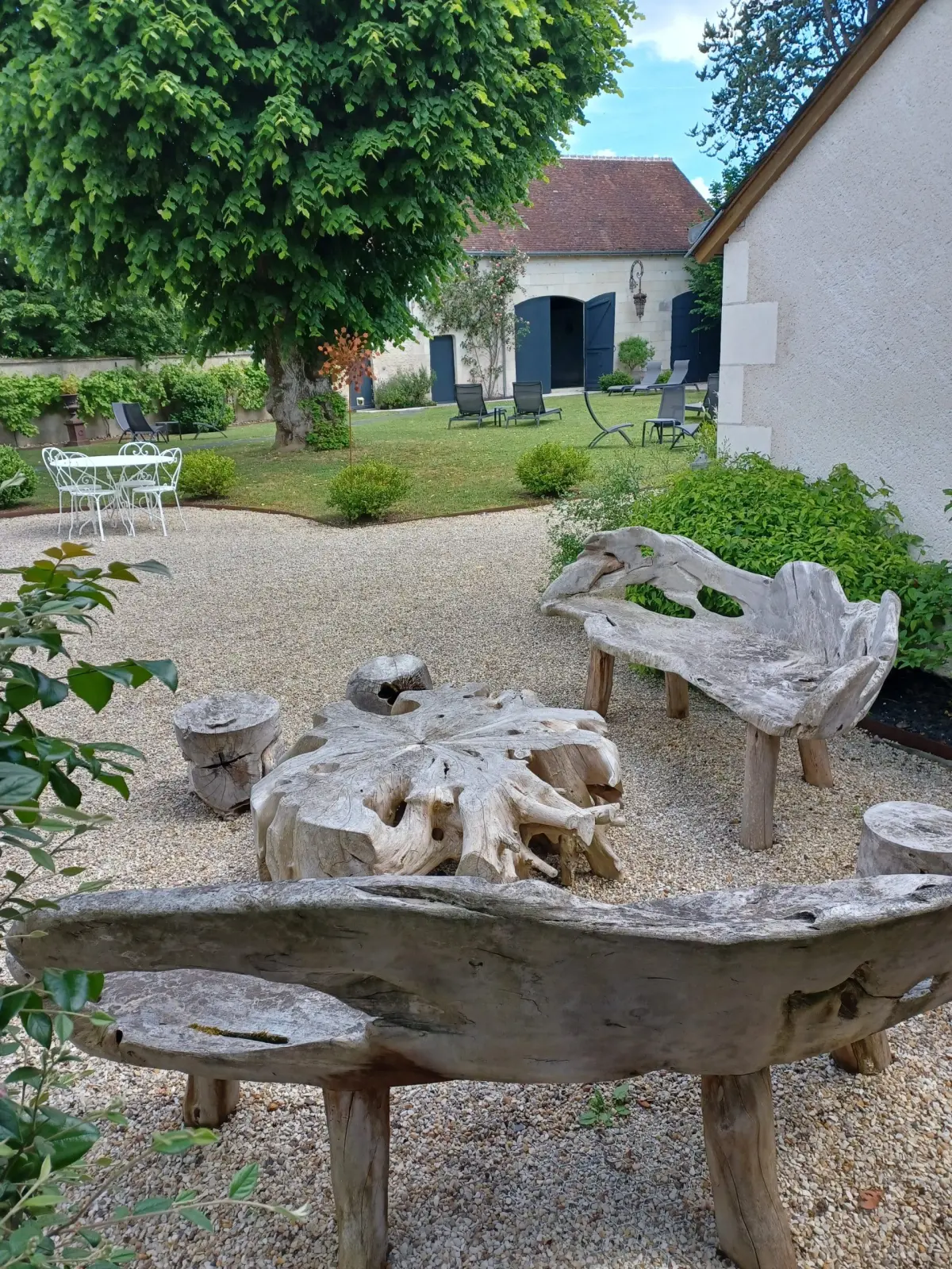 Driftwood benches and table on a gravel courtyard, lawn and loungers behind, La Folie Saint Julien