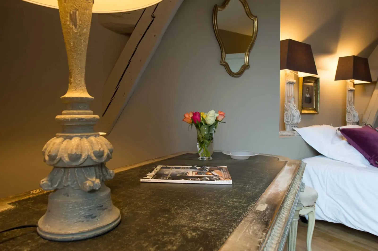 Weathered desk with a carved lamp, a bouquet of roses and a white bed behind, La Folie Saint Julien