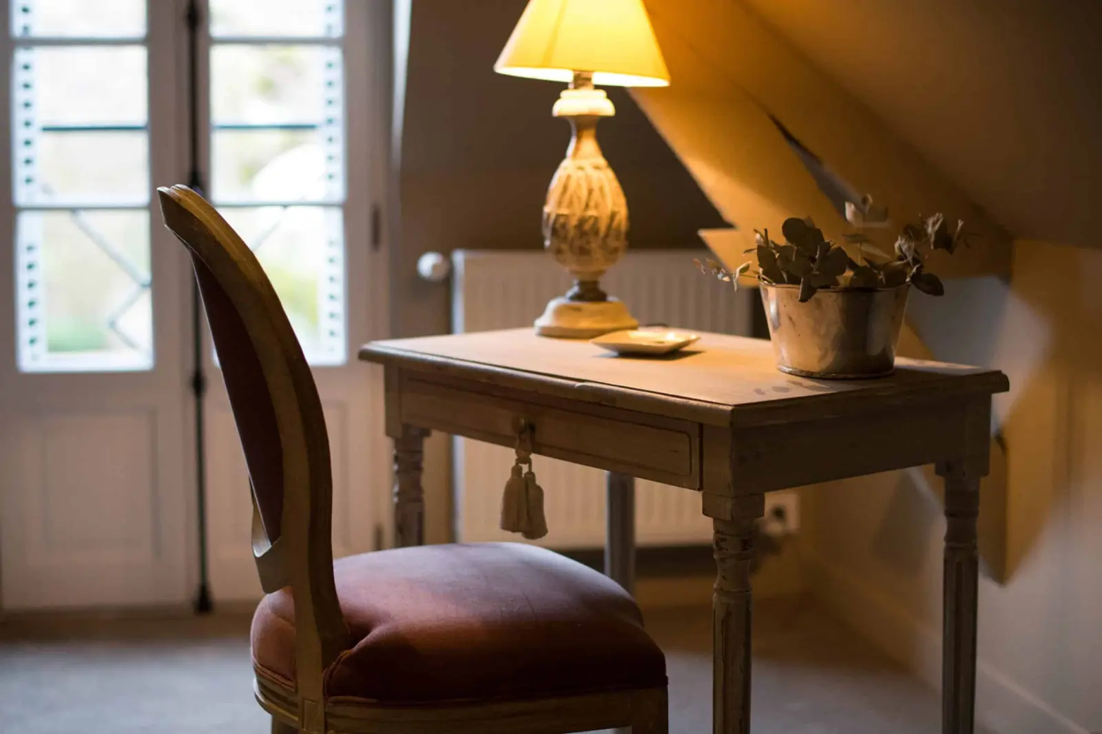 Small weathered desk, velvet chair and a lit lamp in front of a French window, La Folie Saint Julien