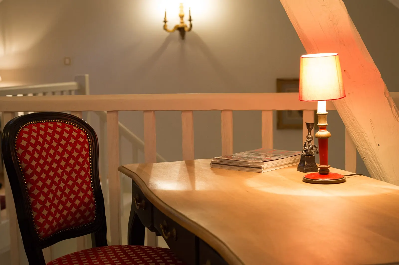 Light wood desk, red patterned chair and a lit lamp by a white banister, La Folie Saint Julien