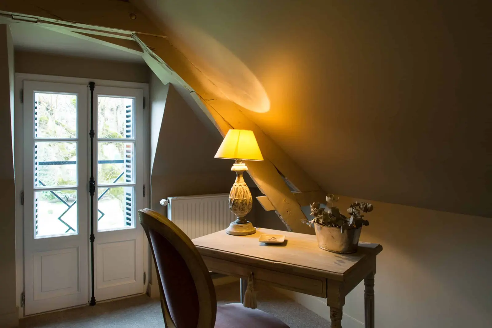 Desk and chair under the sloping roof, a lit lamp by a French window, La Folie Saint Julien