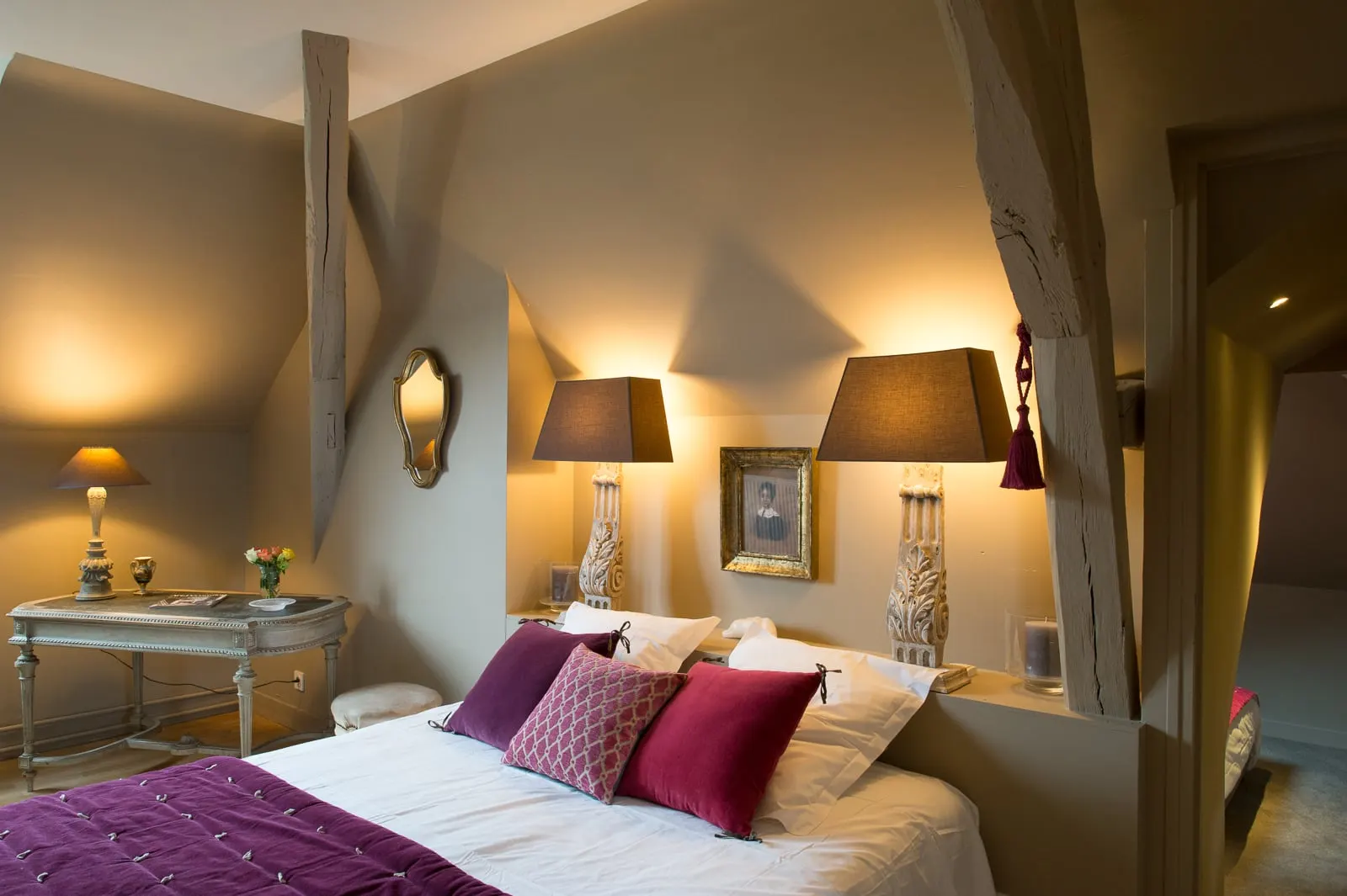 Bedroom with exposed beams, bed with a plum throw, two lamps and a weathered console, La Folie Saint Julien
