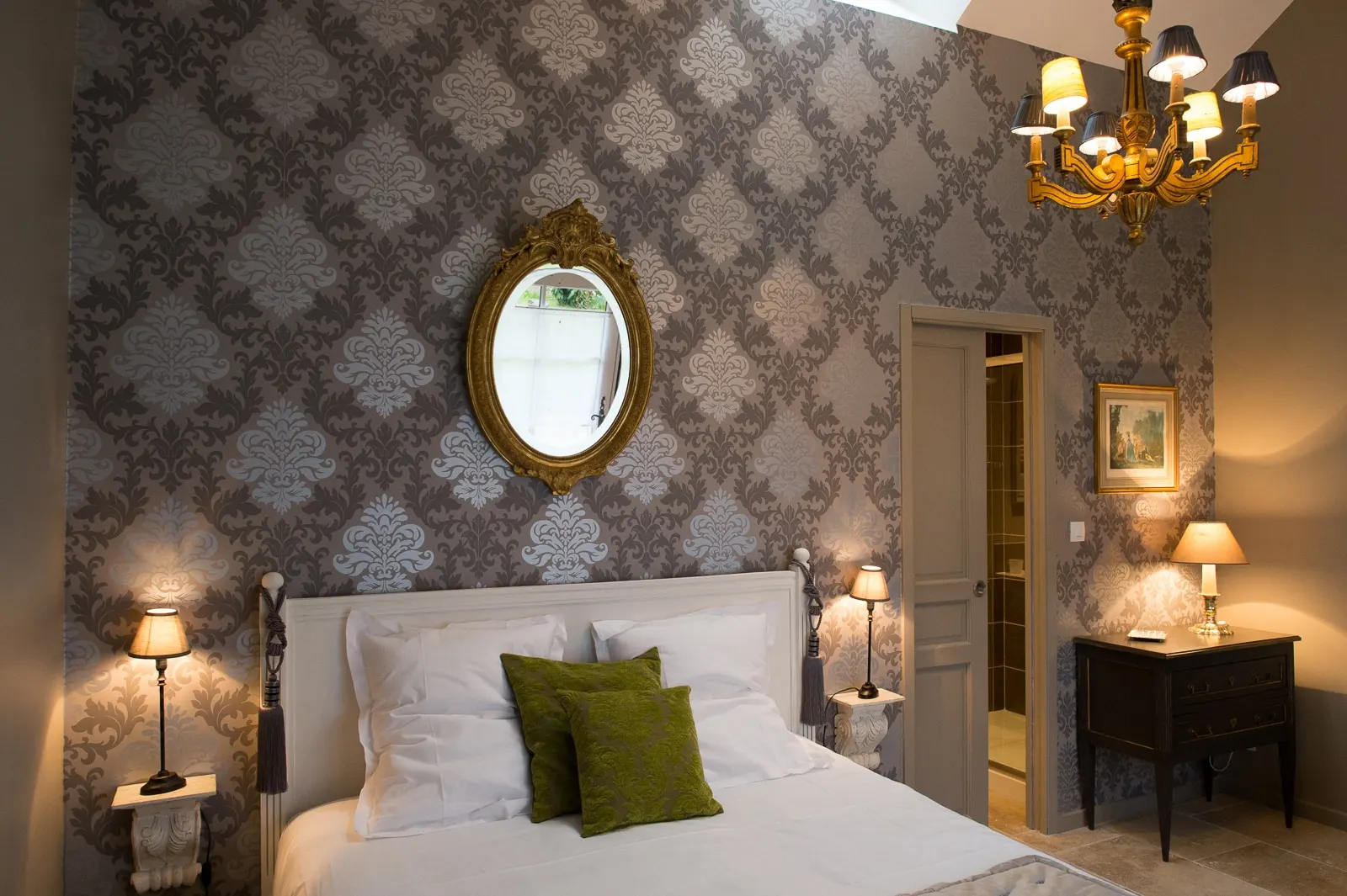 A bedroom at La Folie Saint Julien with a white bed, gilt oval mirror, chandelier and dark chest of drawers