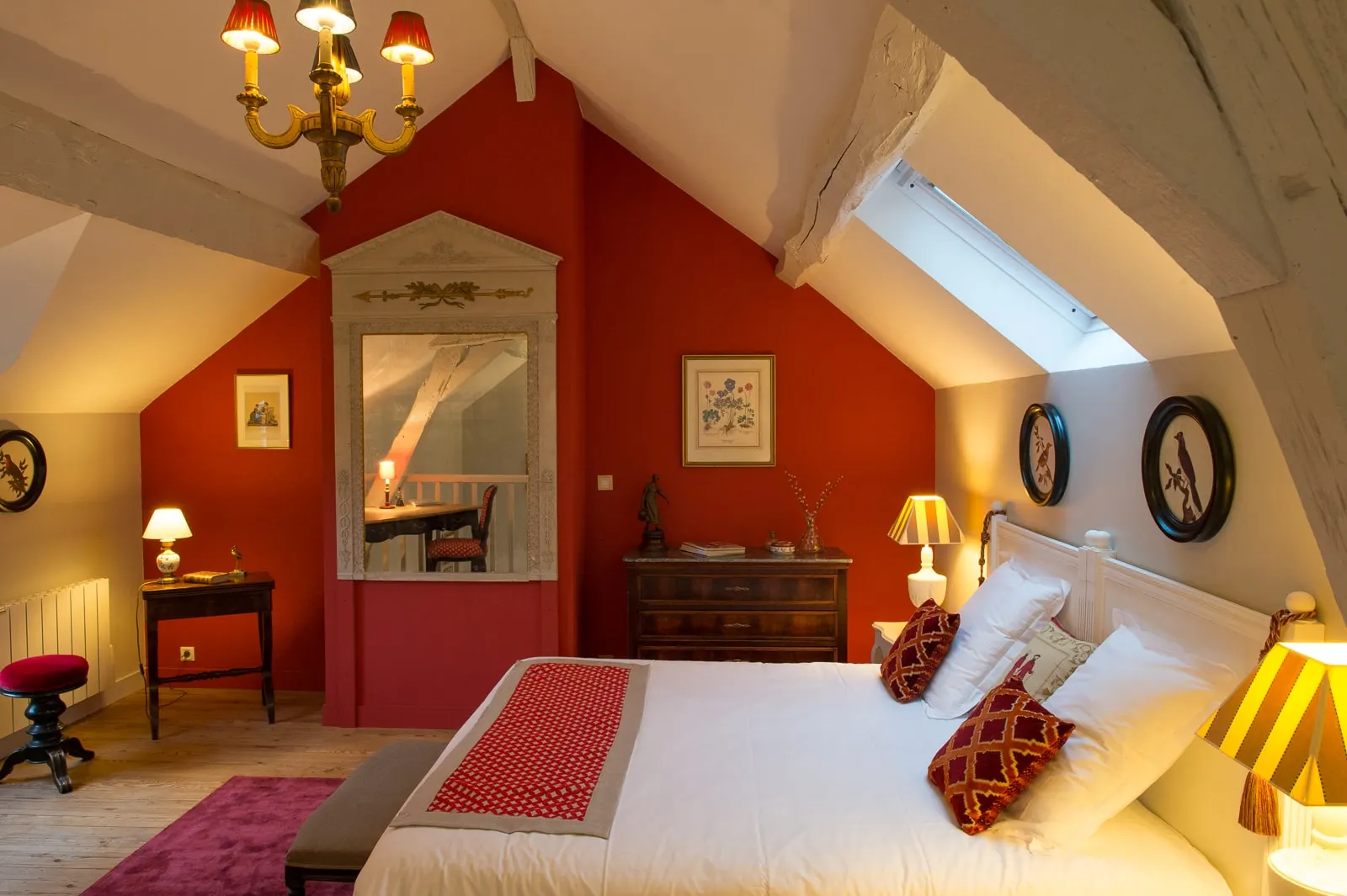 Attic bedroom with a red wall, gilt chandelier, double bed and burgundy cushions, La Folie Saint Julien