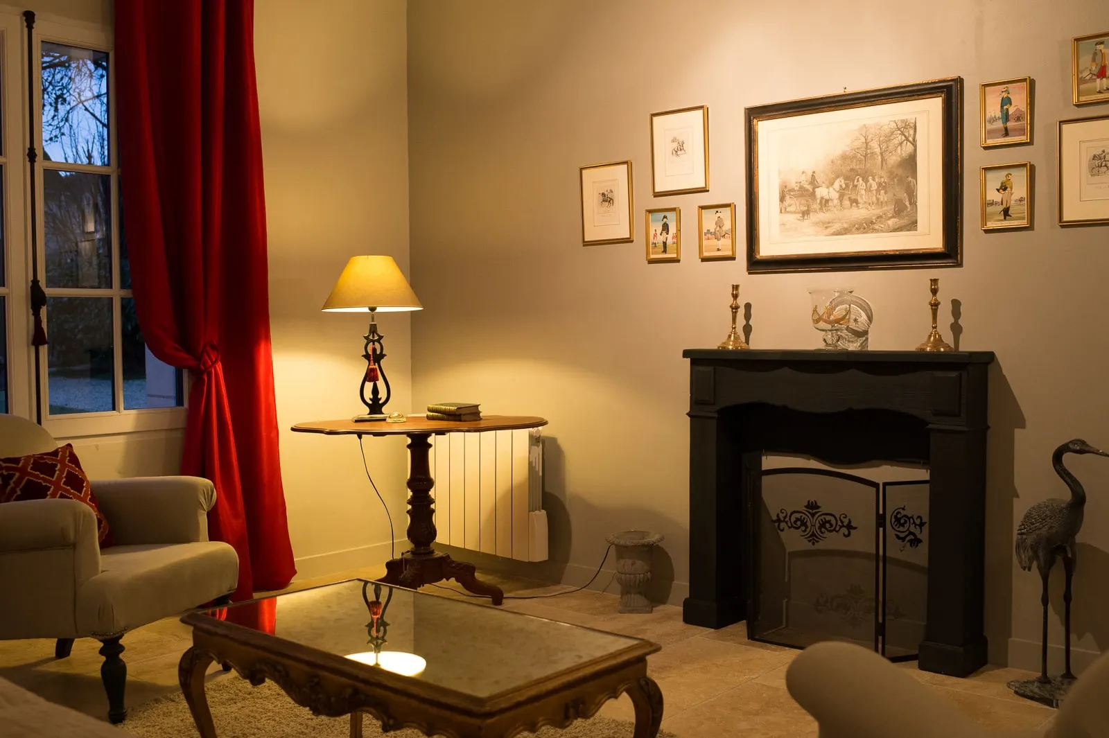 Sitting room in the evening with red curtains, a black fireplace, prints and a lit lamp, La Folie Saint Julien