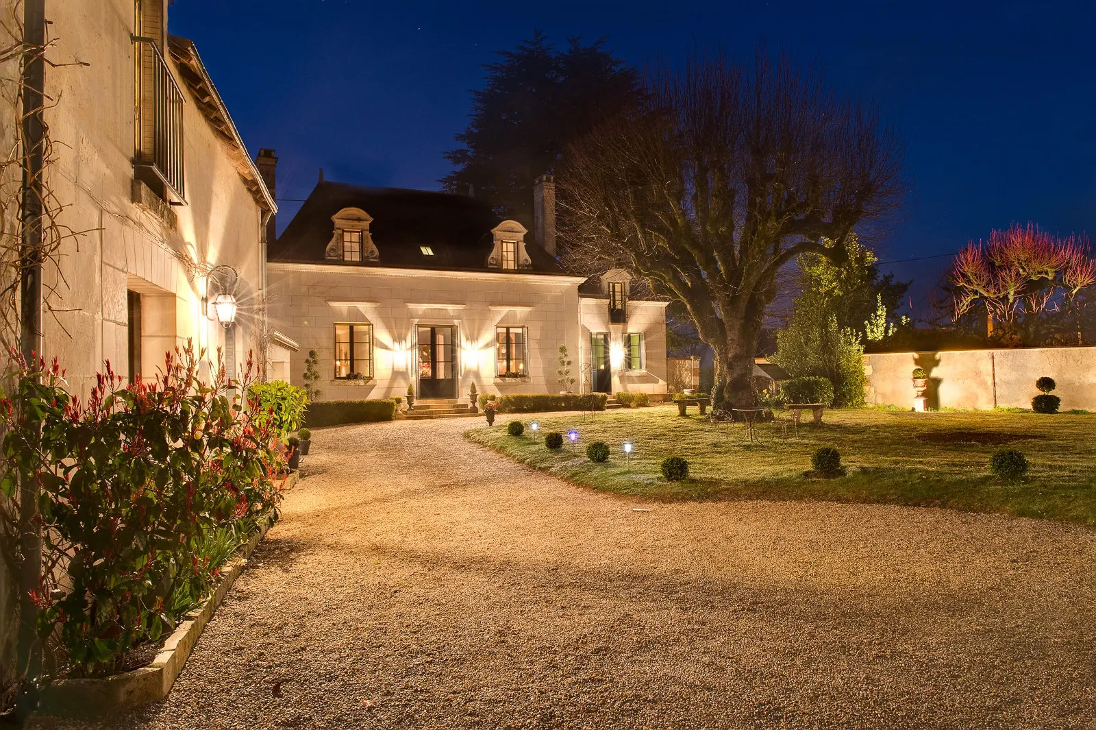 Stone house and gravel courtyard lit up at night, with a lawn and a large tree, La Folie Saint Julien