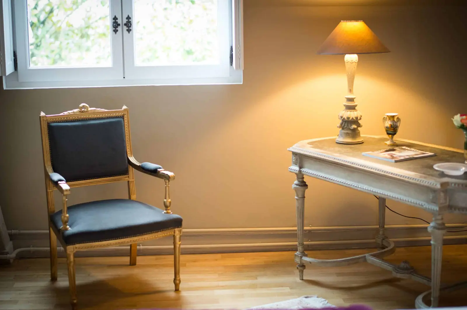 Gilt armchair upholstered in blue beneath a window and a weathered console with a lit lamp, La Folie Saint Julien