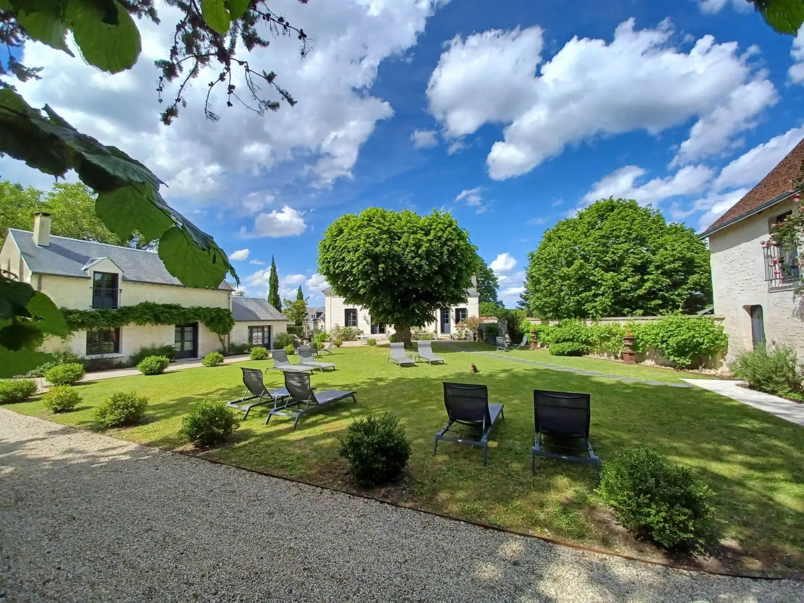 Overview of the garden with loungers, a large tree and stone houses under clouds, La Folie Saint Julien