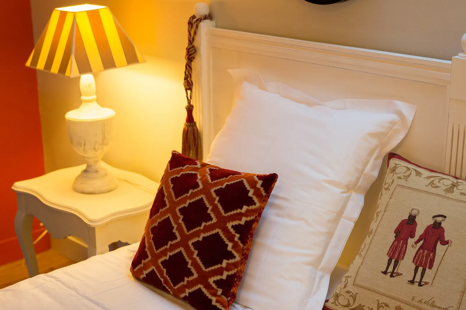 White headboard, embroidered cushions and a striped-shade lamp on the bedside table, La Folie Saint Julien