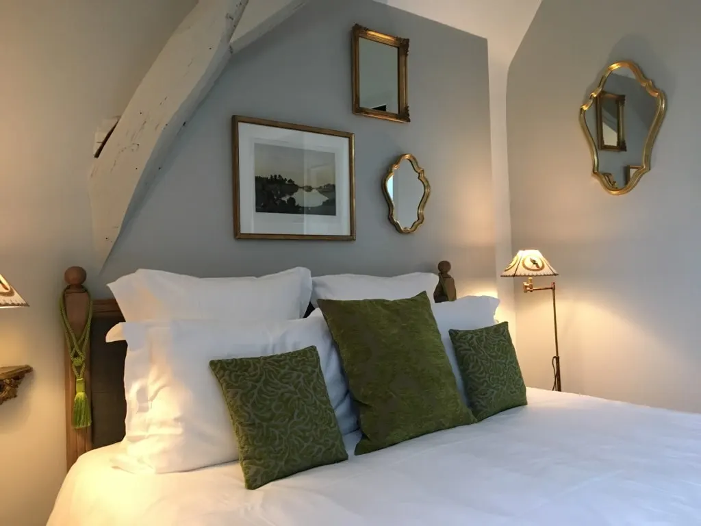 White bed with olive-green cushions under a beam, gilt mirrors and a print, La Folie Saint Julien