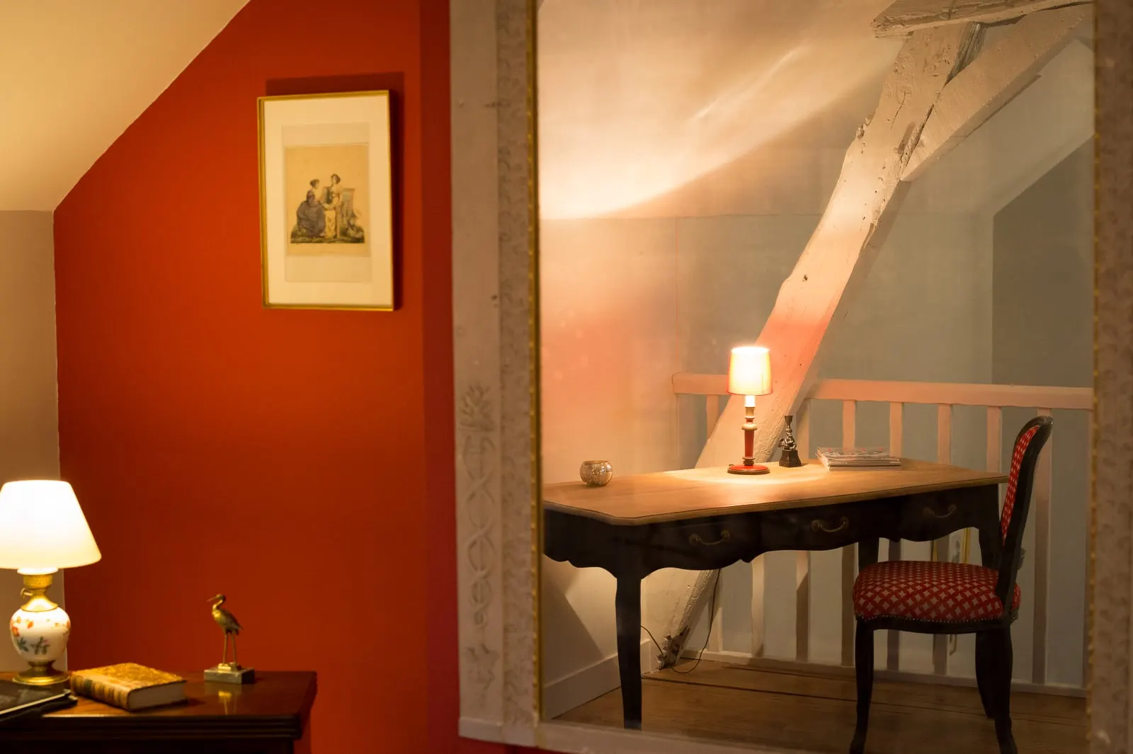 Mirror reflecting a desk and its lit lamp under the beams, beside a red wall, La Folie Saint Julien