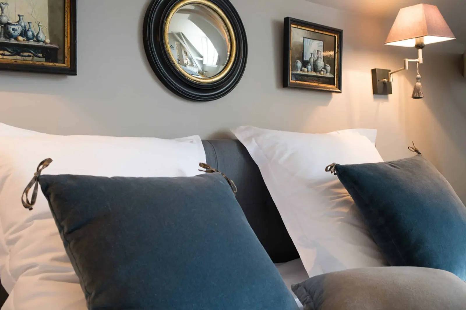 Blue velvet cushions on white pillows, a round mirror and a lit wall lamp, La Folie Saint Julien
