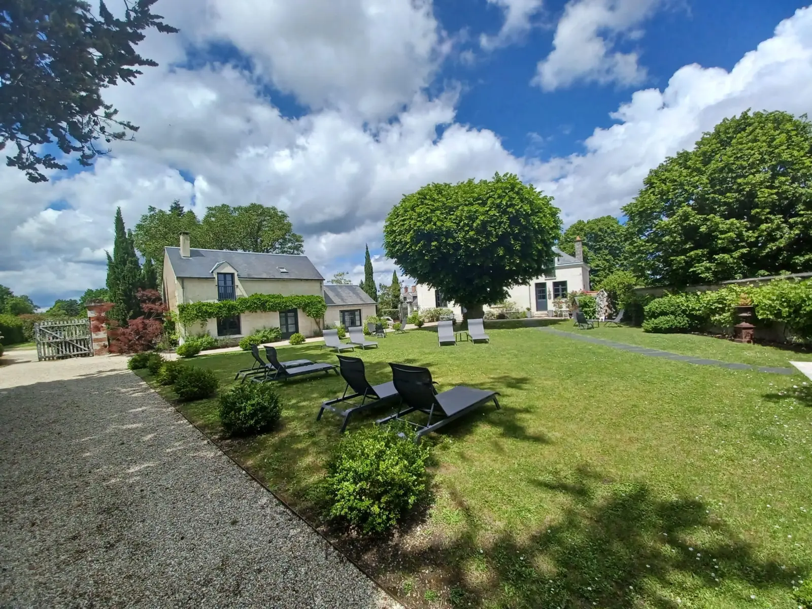 Sunny lawn with black loungers, trees and stone houses under a cloudy sky, La Folie Saint Julien