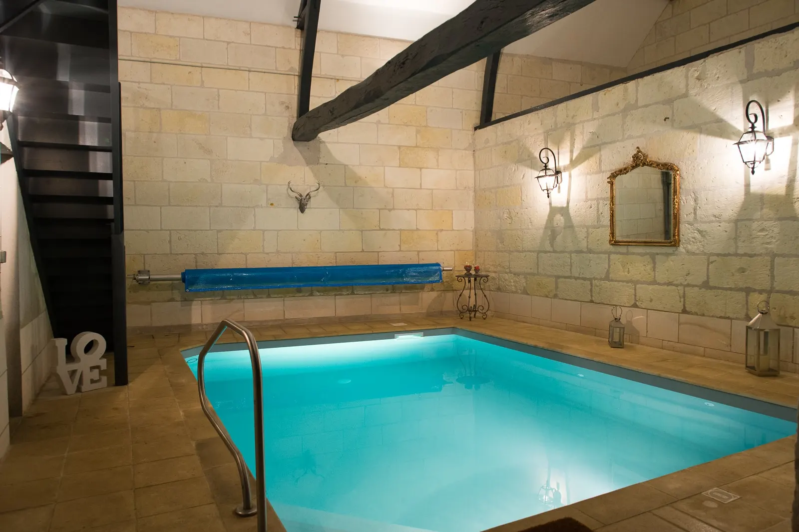 Lit indoor pool between stone walls and dark beams, with a black staircase, La Folie Saint Julien