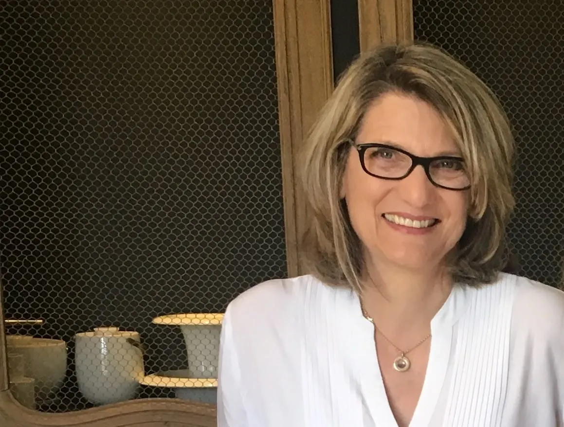 Smiling host in a white blouse and glasses in front of a wire-mesh cabinet, La Folie Saint Julien