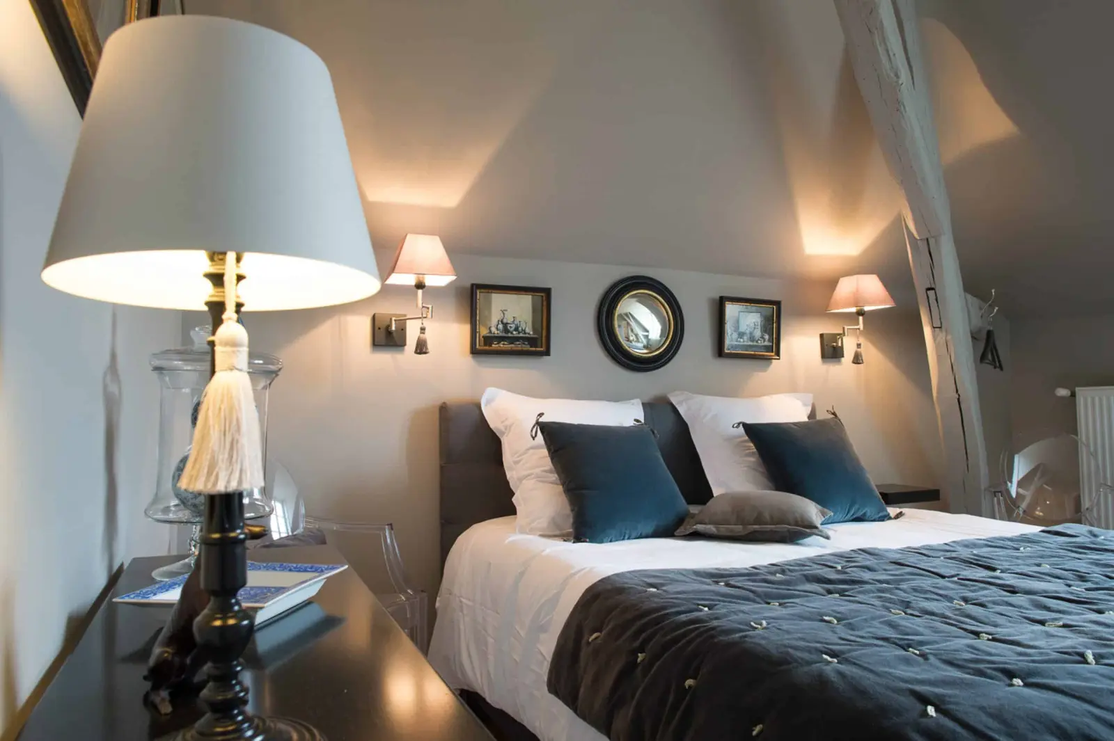 Attic bedroom in grey and blue tones, double bed, wall lamps and a round mirror, La Folie Saint Julien
