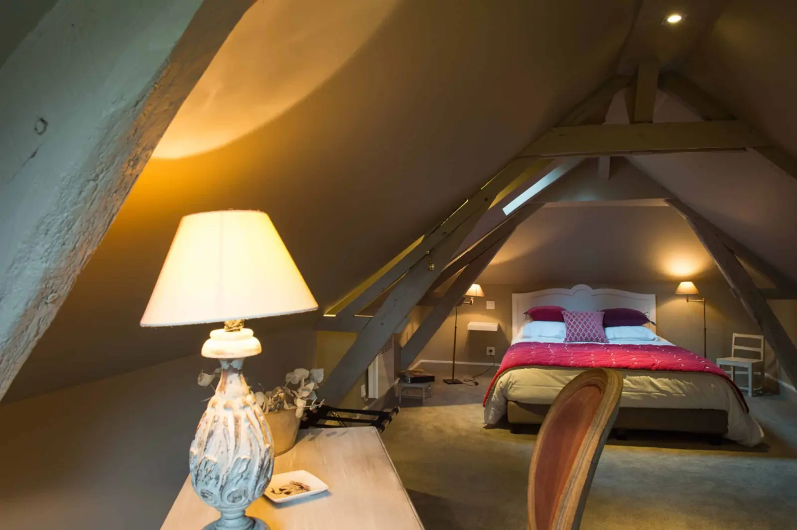 Large bedroom beneath the roof timbers, bed with a raspberry throw and a desk with a lit lamp, La Folie Saint Julien