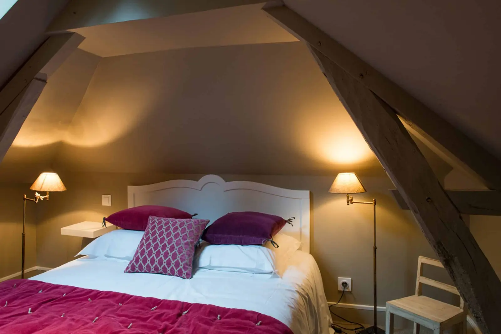 Attic bedroom beneath the roof timbers, white headboard, plum cushions and reading lamps, La Folie Saint Julien