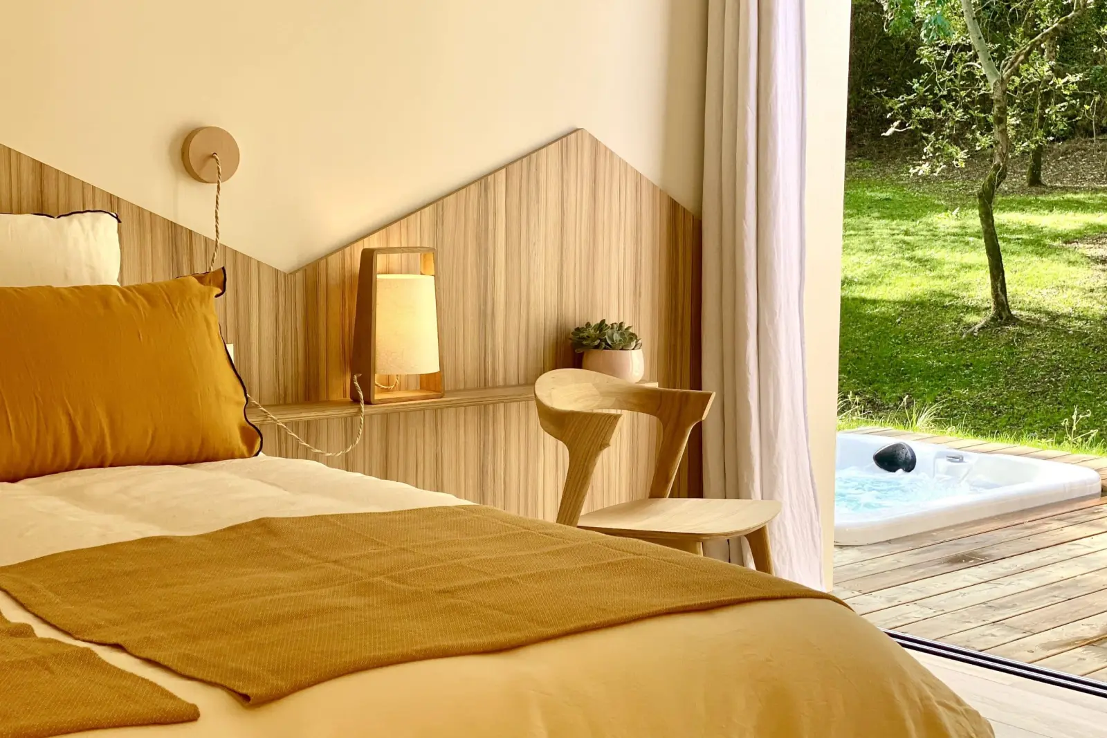 Bedroom at Kassiopée: bed with mustard linen, wooden headboard and glass door open onto the outdoor hot tub
