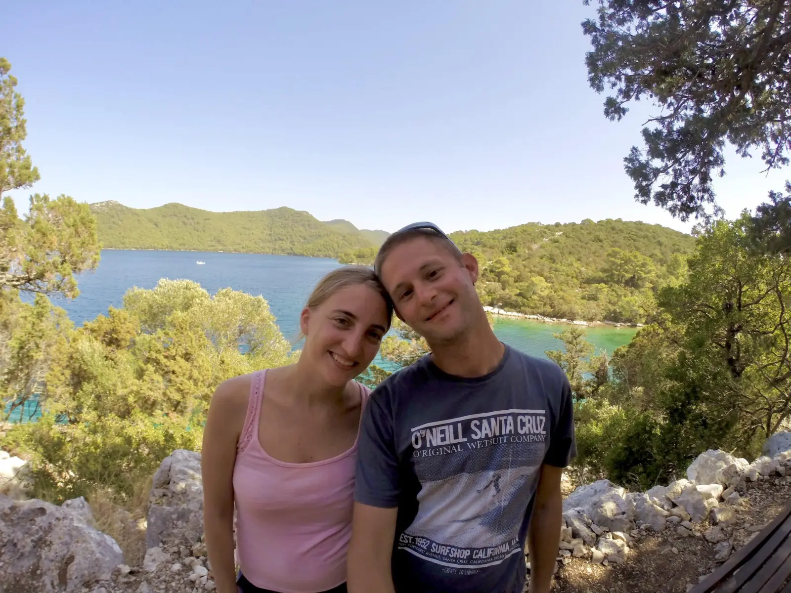 The Kassiopée host couple smiling in front of a turquoise bay and wooded hills
