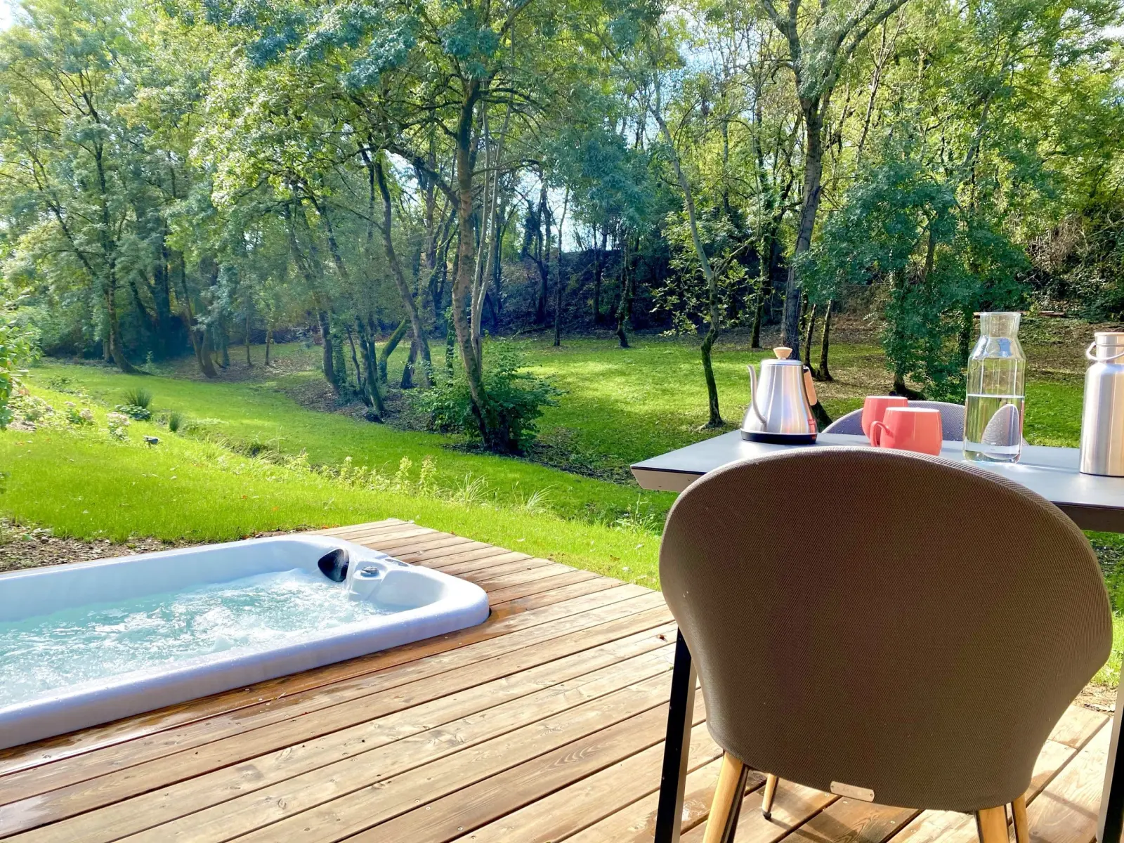 Spa privatif encastré dans une terrasse en bois de Kassiopée, table avec théière et vue sur le parc