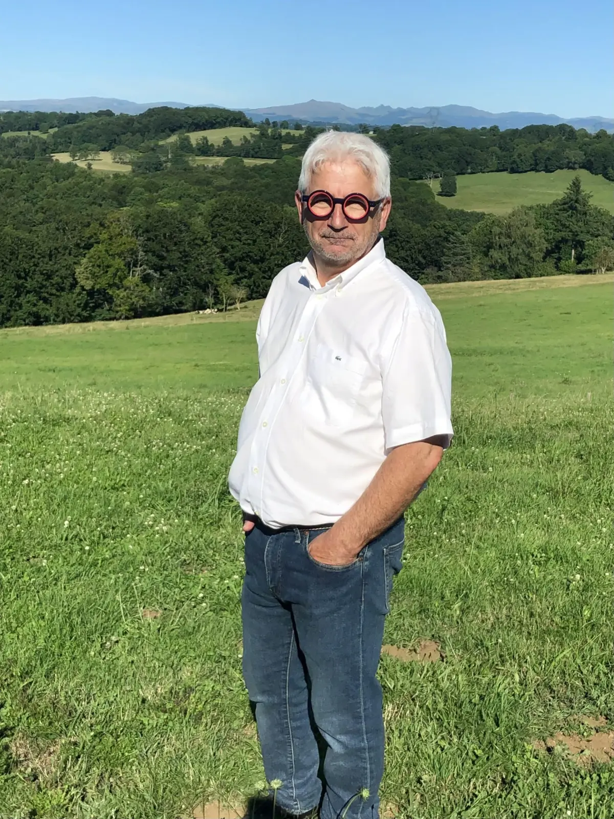 White-haired man in a white shirt standing in a meadow facing the hills, La Fournio