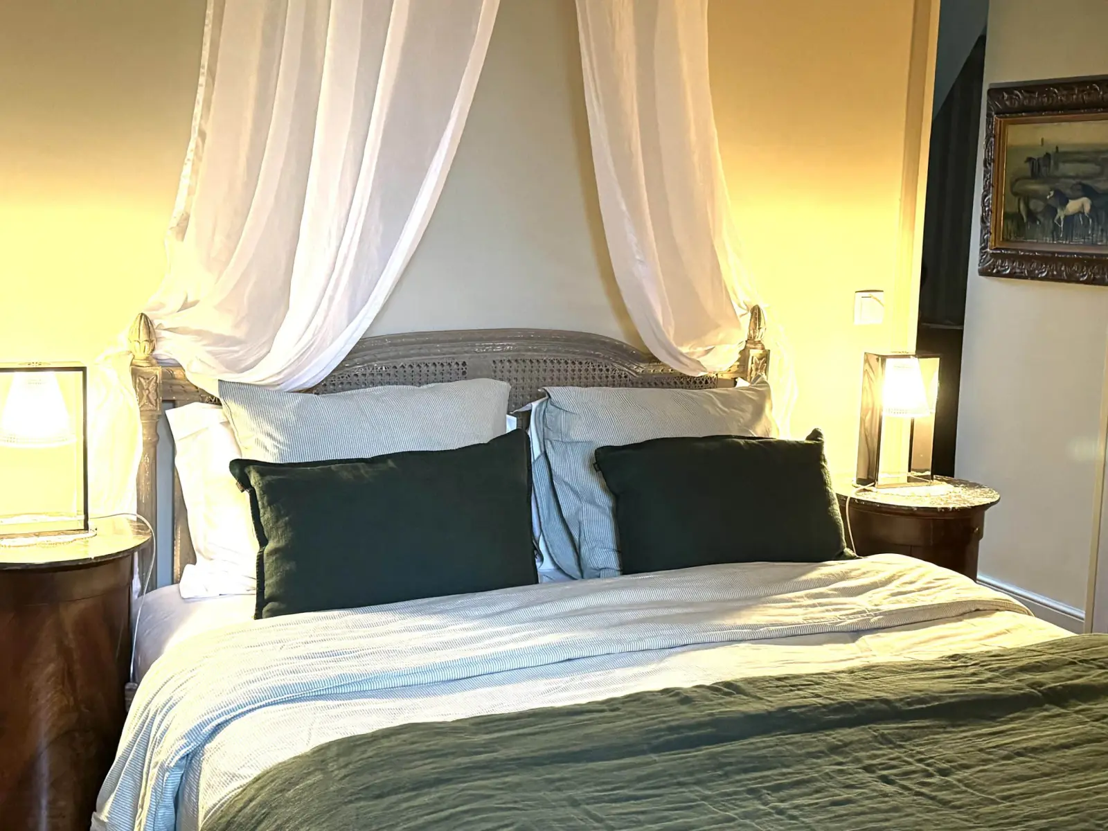 Double bed beneath a white voile canopy, dark green cushions and wooden bedside tables, La Fournio