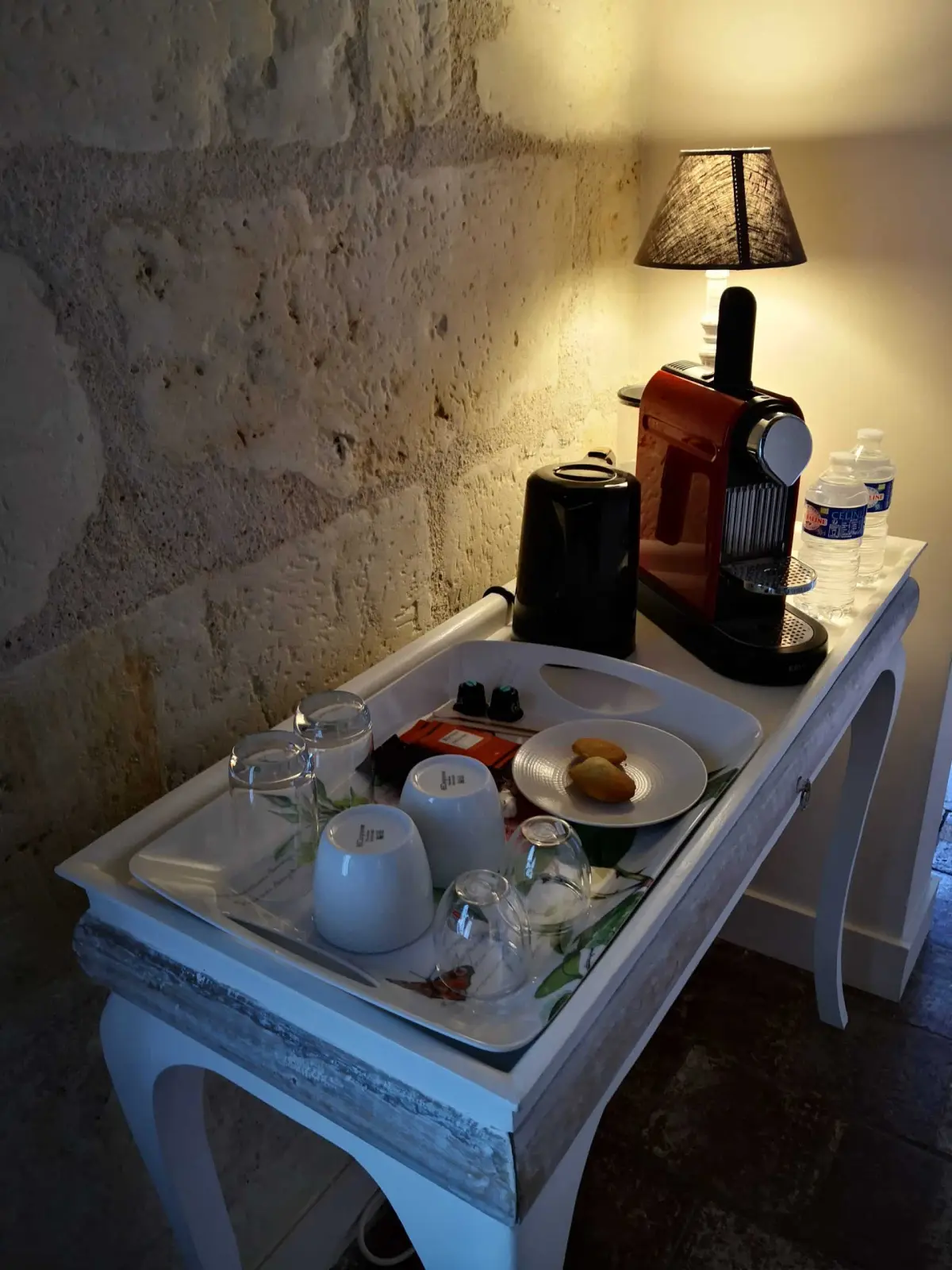 Welcome tray with coffee machine, cups, glasses and biscuits on a white console against a stone wall, La Vie Voyage