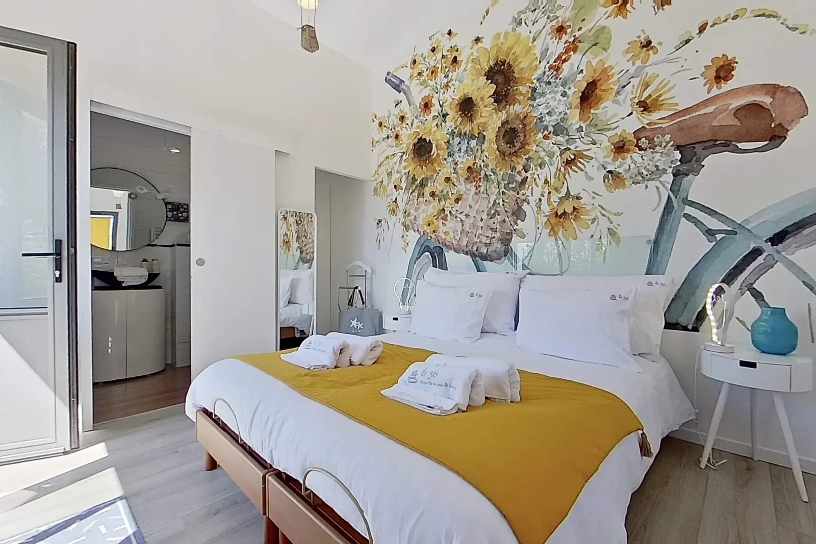 Bedroom at Le 36 with double bed, yellow throw, towels and a mural of a bicycle with a basket of sunflowers